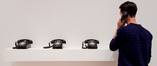 a row of black, analog telephones in a white room. person facing the row of phones, with receiver lifted to left ear.